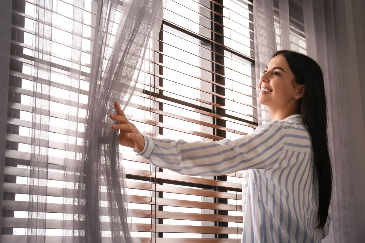 the-benefits-of-a-professional-window-treatment-consultation-1200px Smiling woman standing in front of a window with custom blinds and drapery at The Blind Guy near Spearfish, SD